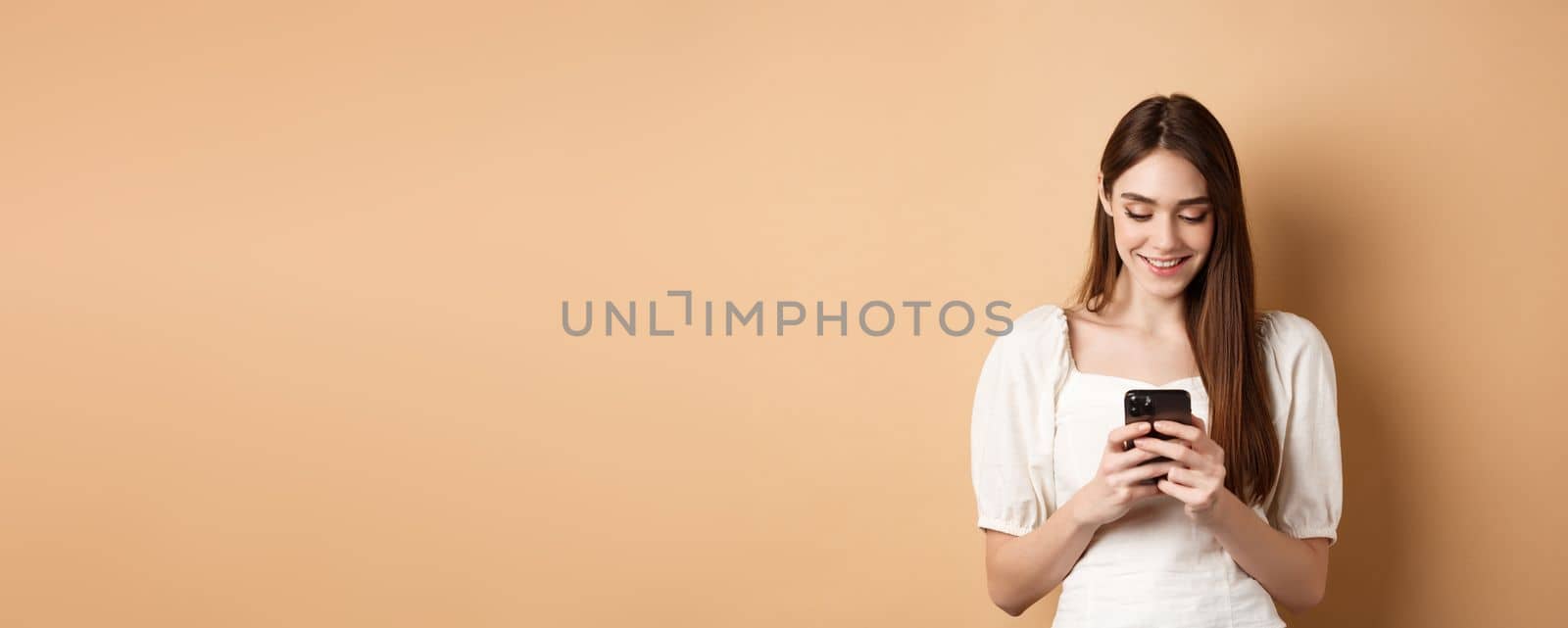Young woman texting message on mobile phone, smiling and reading smartphone screen, standing on beige background.