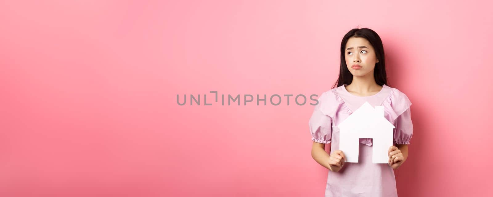 Real estate and insurance concept. Sad asian woman looking aside with unfair face, showing paper house cutout, dreaming of apartment, standing against pink background.