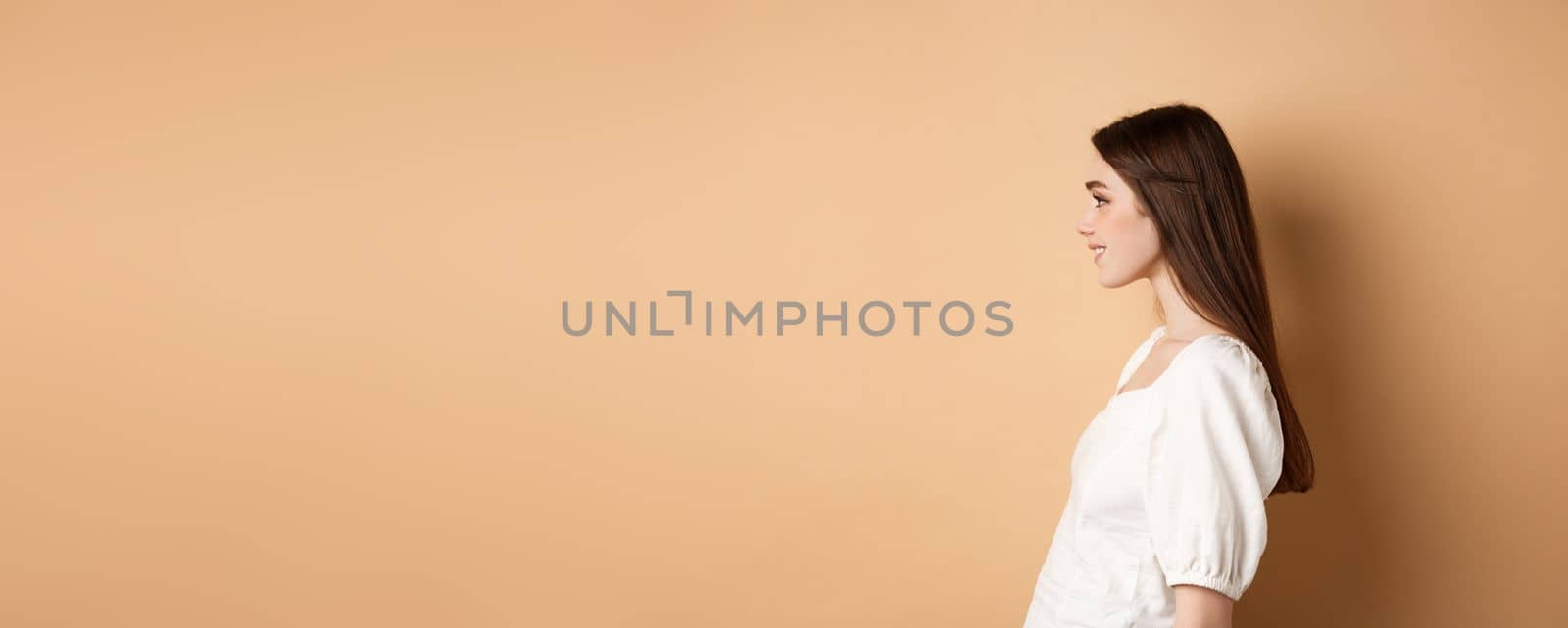 Profile of beautiful caucasian woman with long natural hair, looking left at empty space and smiling romantic, standing on beige background by Benzoix