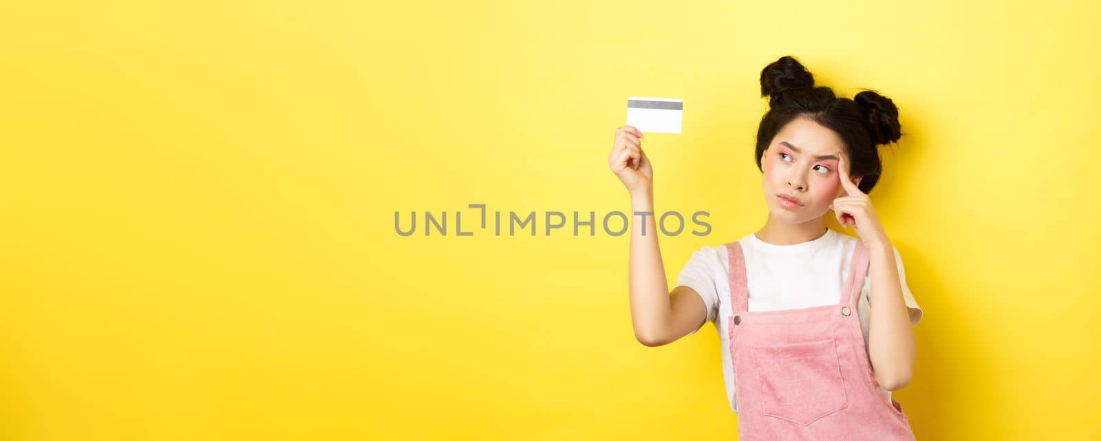 Shopping. Thoughtful trendy girl with summer makeup, looking pensive at plastic credit card, standing on yellow background by Benzoix
