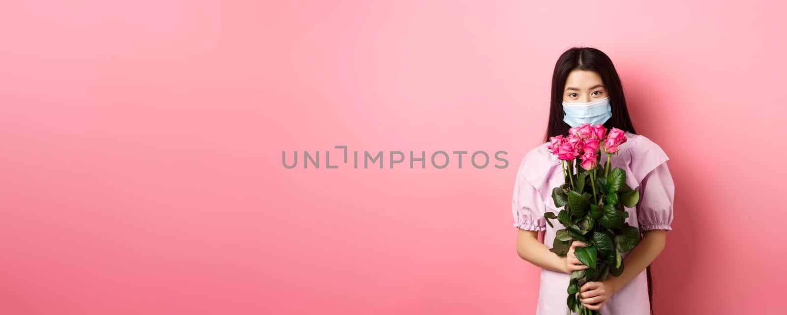 Young asian girl in medical mask holding flowers on Valentines day, receive bouquet of roses from lover, standing on pink background. Social distancing and covid-19 concept by Benzoix