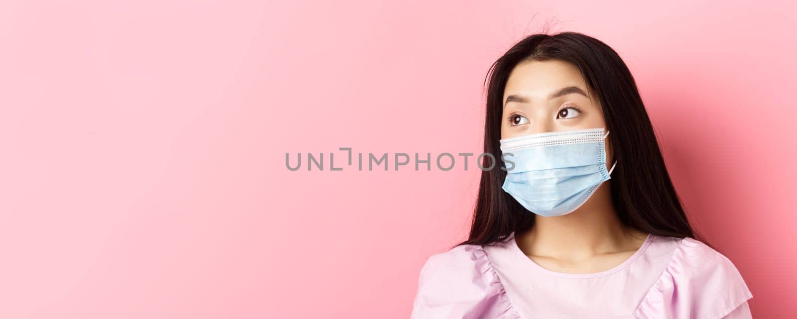 Covid-19, pandemic and quarantine concept. Close-up of dreamy asian girl in medical mask looking left at logo with thoughtful face, standing on pink background.