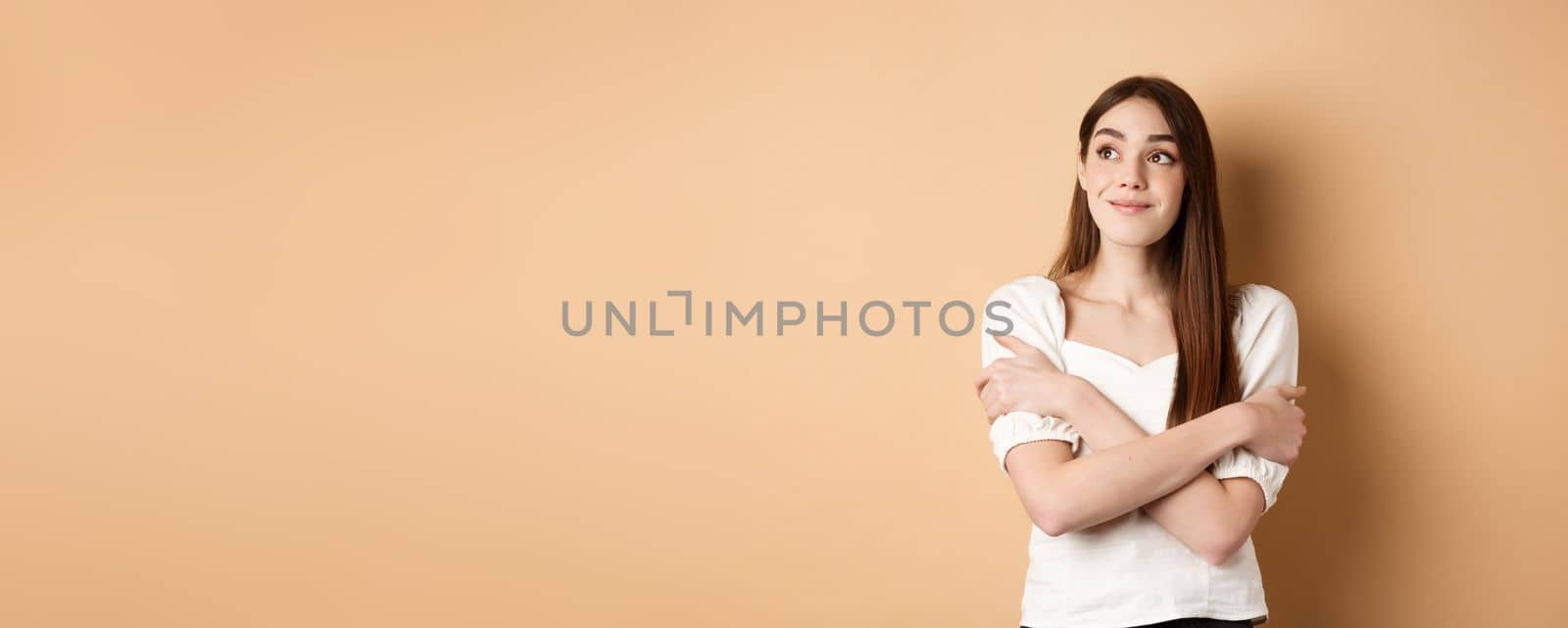 Romantic young girl dreaming, hugging herself and smiling at upper left corner, imaging something lovely, standing on beige background.