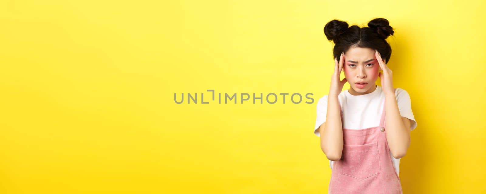 Image of stylish japanese woman with bright makeup, having headache, touching head and looking tired, have painful migraine, yellow background by Benzoix