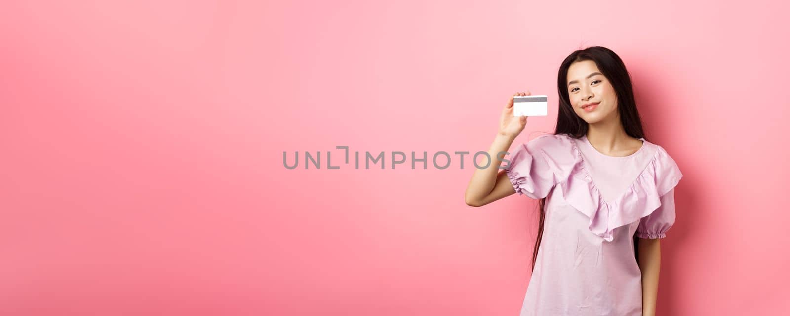 Shopping. Young korean girl in dress showing credit card, smiling satisfied at camera, standing on pink background by Benzoix