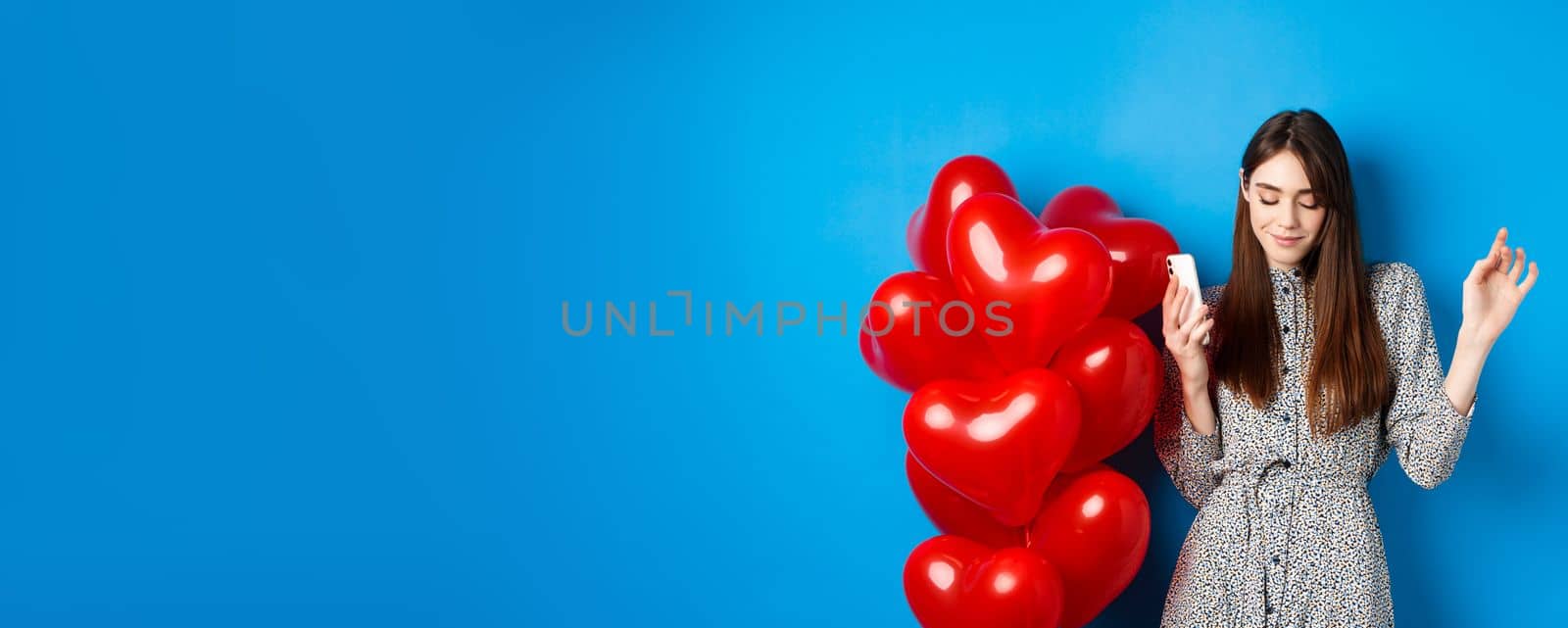 Portrait of attractive young woman holding smartphone and dancing near Valentines red balloons, standing on blue background by Benzoix