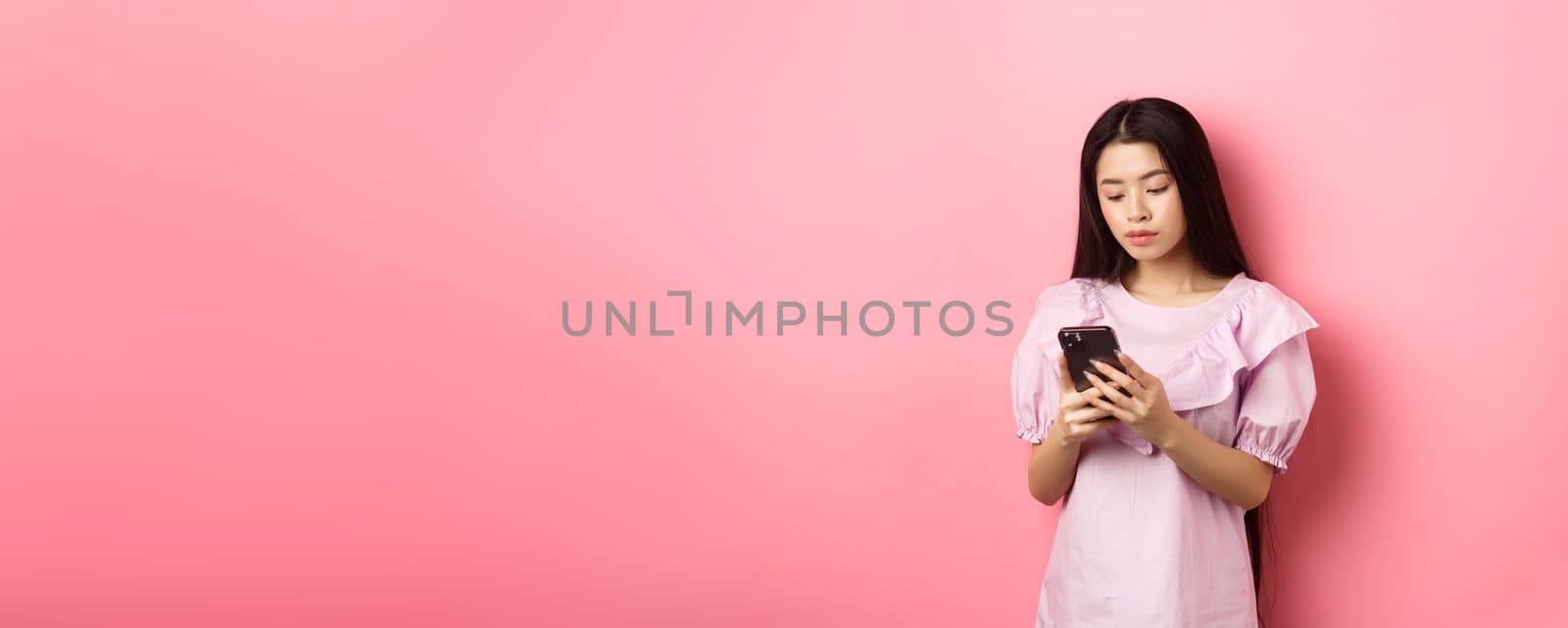 Teenage asian girl chatting on mobile phone, look at smartphone screen with serious face, standing against pink background by Benzoix