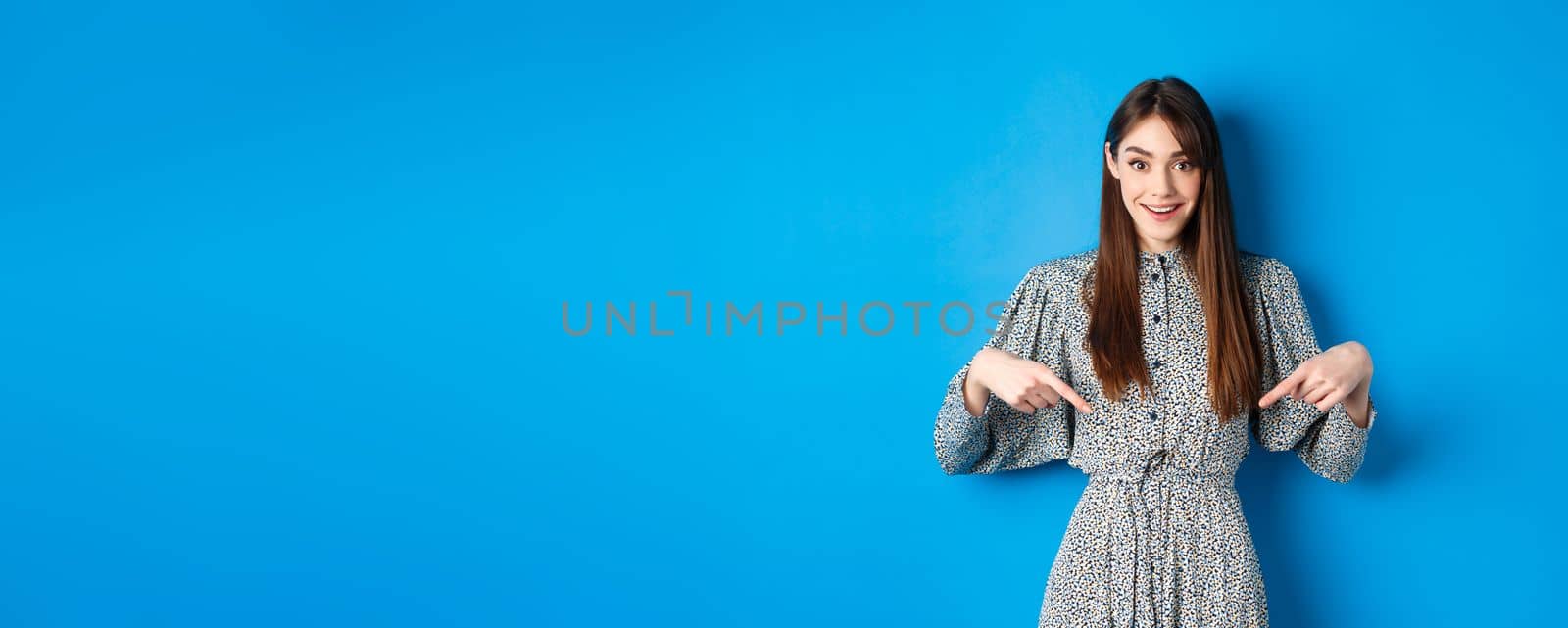Excited caucasian woman in dress pointing fingers down at logo, showing fantastic news and smiling, standing on blue background by Benzoix