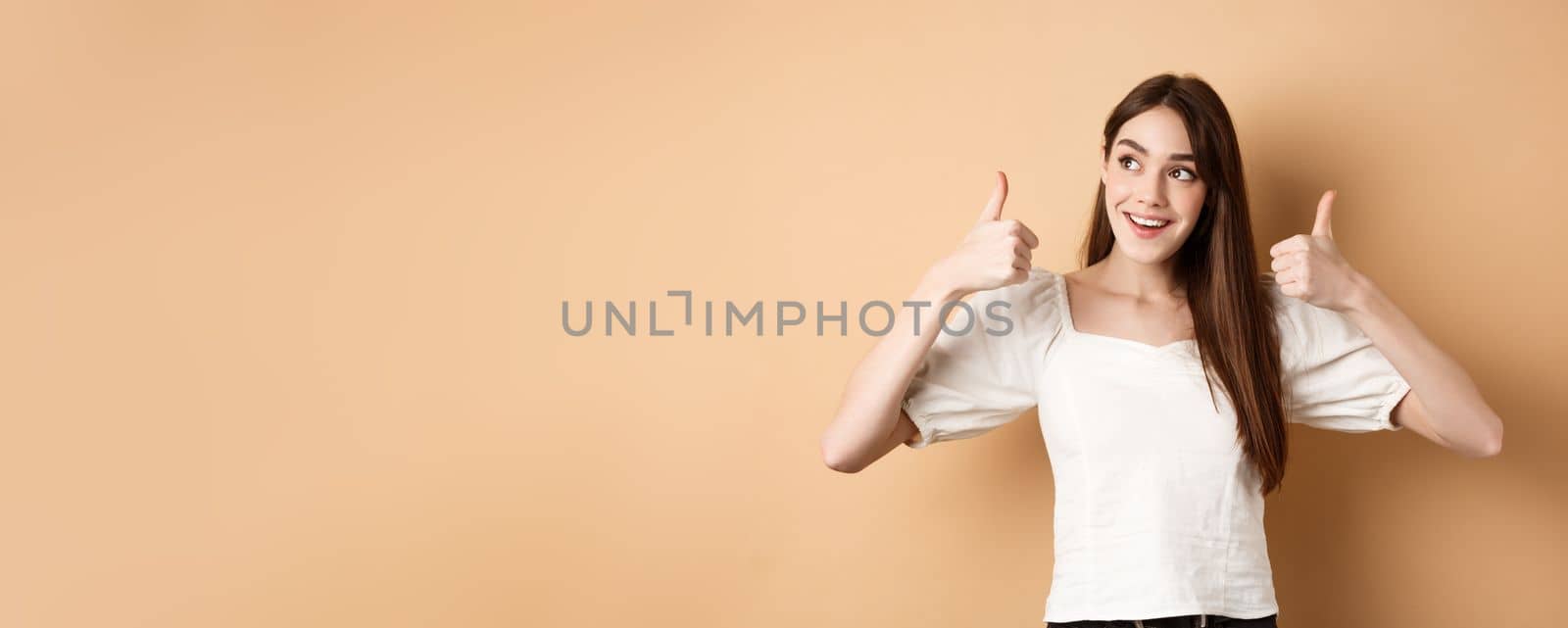 Smiling dreamy girl looking at upper left corner and showing thumbs up at promo, recommending product, praising banner, standing on beige background by Benzoix