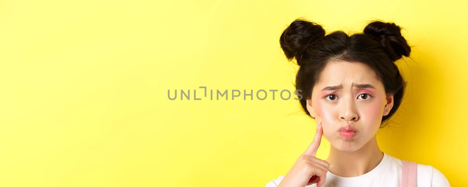 Close-up portrait of stylish asian girl with bright makeup and glowing skin, pouting and frowning, poking her cheek with sad face, standing upset on yellow background.