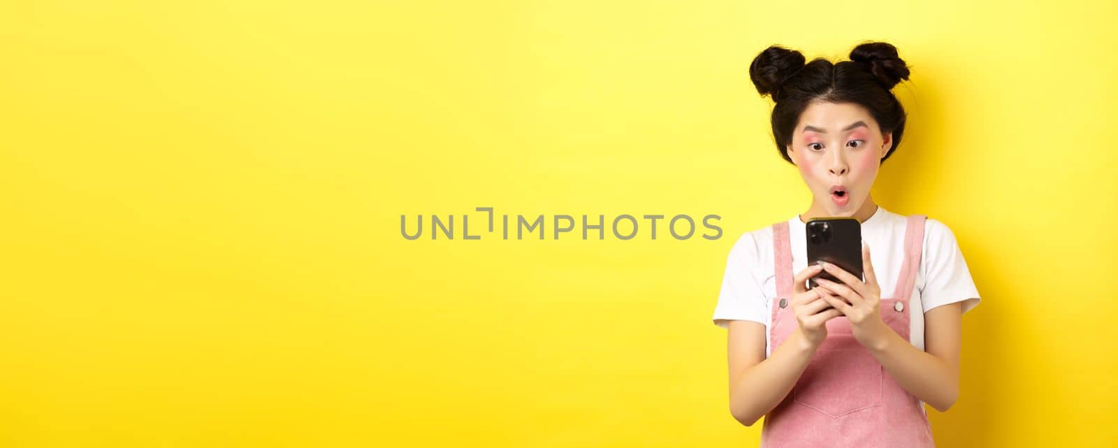 Asian teen girl look excited at smartphone screen, reading news on phone, standing on yellow background.