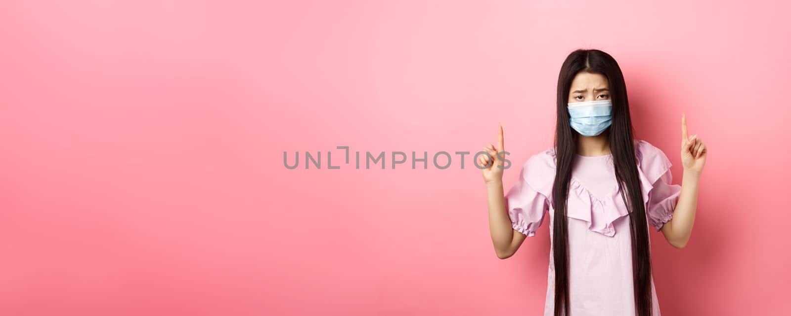 Healthy people and covid-19 pandemic concept. Sad asian girl feeling sick, wearing medical mask on self-quarantine, standing against pink background by Benzoix