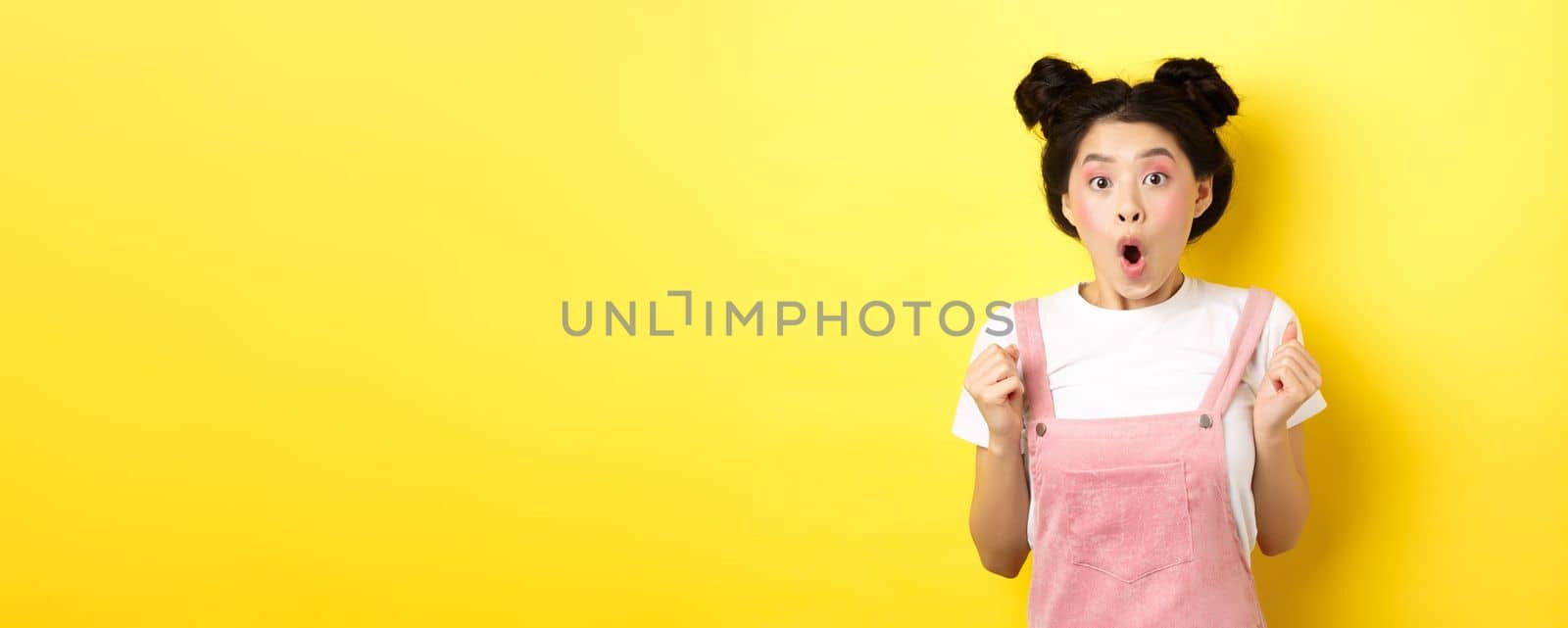 Excited and surprised asian girl clench fists, looking hopeful and amazed, say wow, standing on yellow background by Benzoix