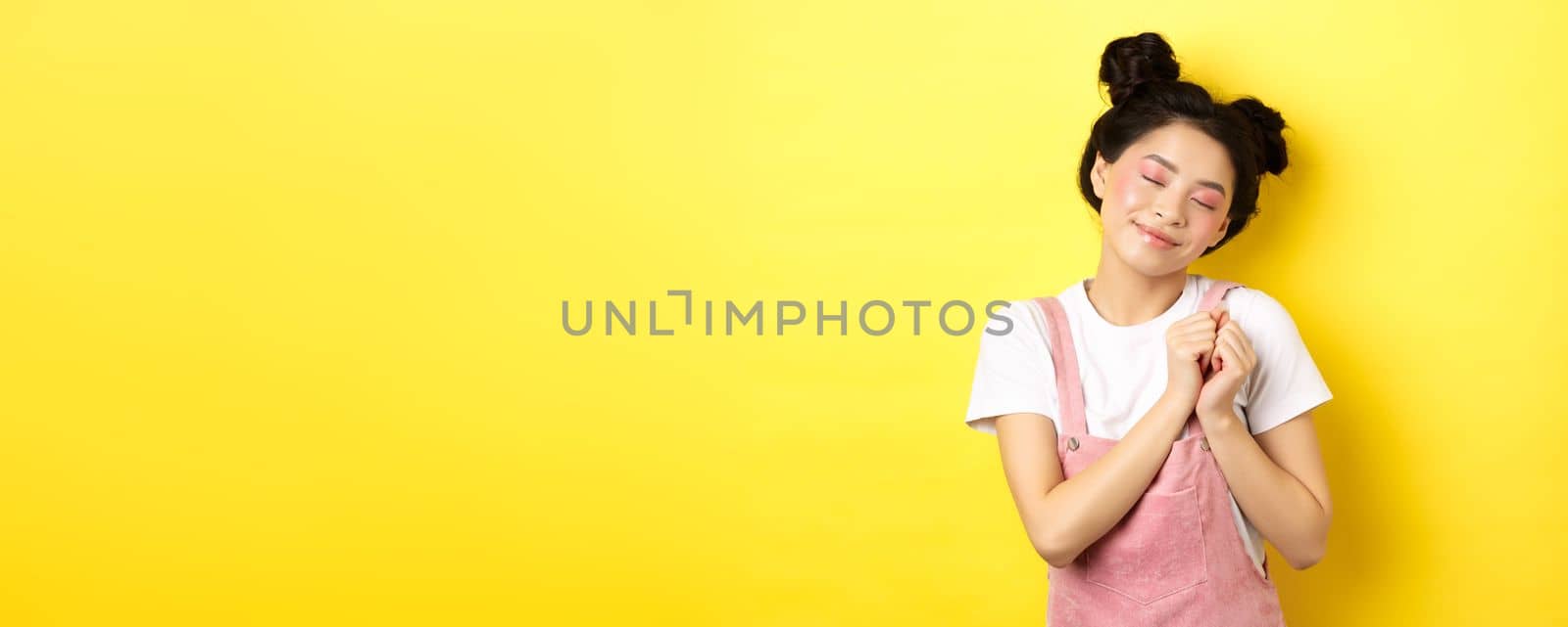 Cute asian girl with makeup, close eyes and remember beautiful moment, holding hands on heart daydreaming, standing on yellow background.