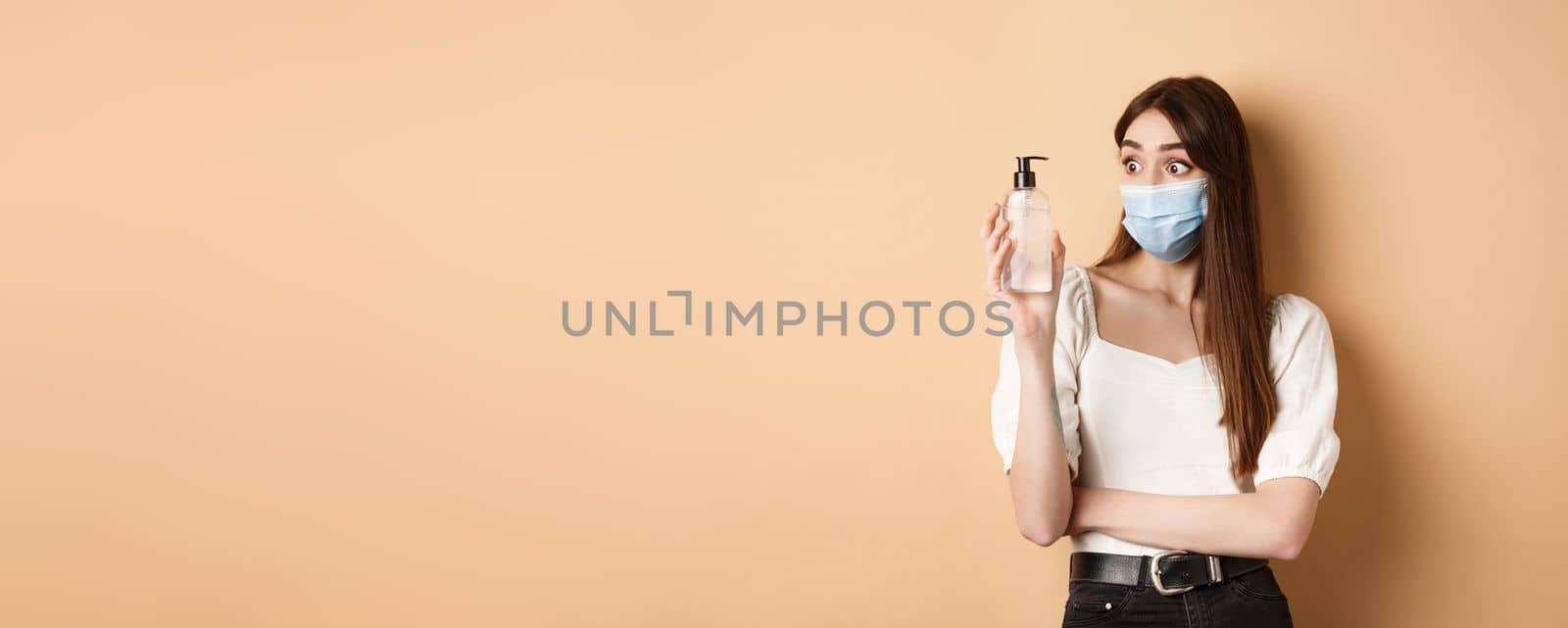 Covid-19 and preventive measures concept. Excited girl in medical mask look amazed at hand sanitizer bottle, standing on beige background.