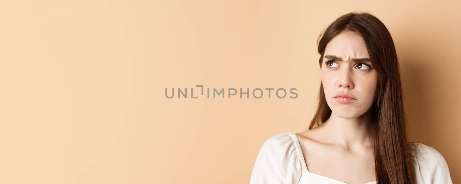 Close up of angry cute girl frowning, looking aside displeased, stare at bad thing, standing on beige background.