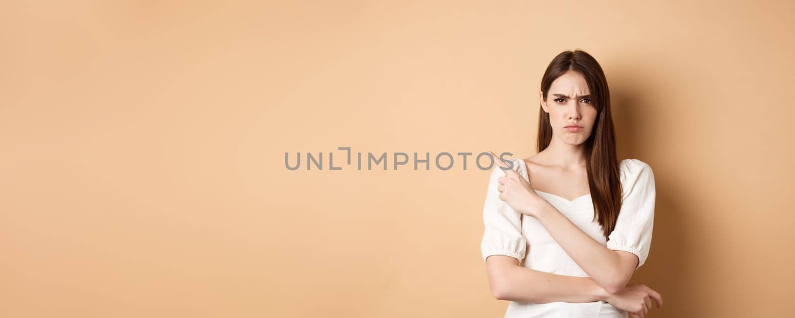 Angry frowning girl pointing left at empty space, looking disappointed and upset, scolding you or accuse, standing on beige background.