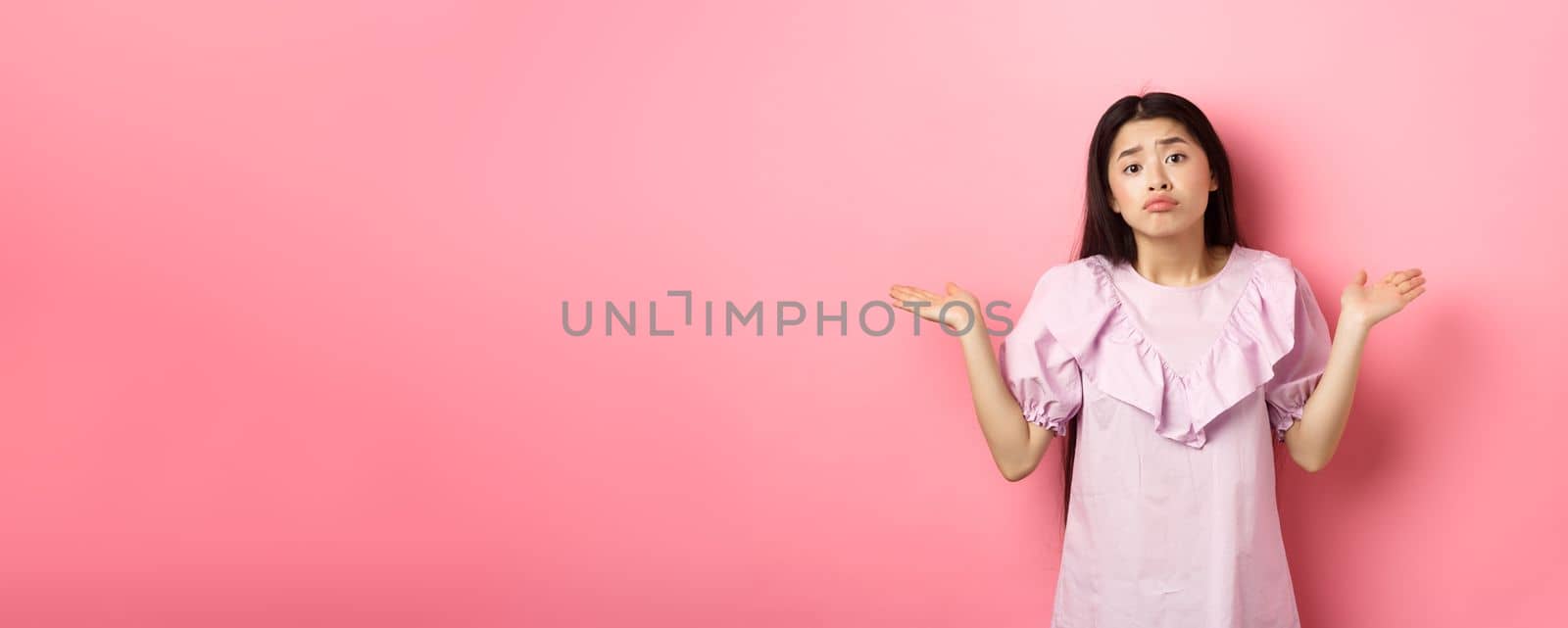 Sorry cant help. Clueless asian girl shrugging with sad face, know nothing, standing indecisive against pink background by Benzoix