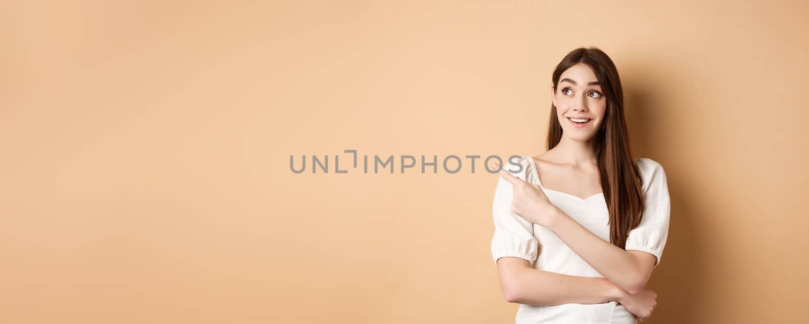 Cheerful tender girl in white dress checking out promo offer, looking and pointing at upper left corner with dreamy smile, standing on beige background.