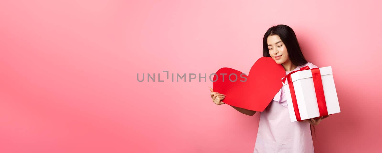 Valentines day concept. Romantic asian teen girl receive gifts on white day from lover, hugging big red heart card and present in box, smiling sensual, pink background by Benzoix