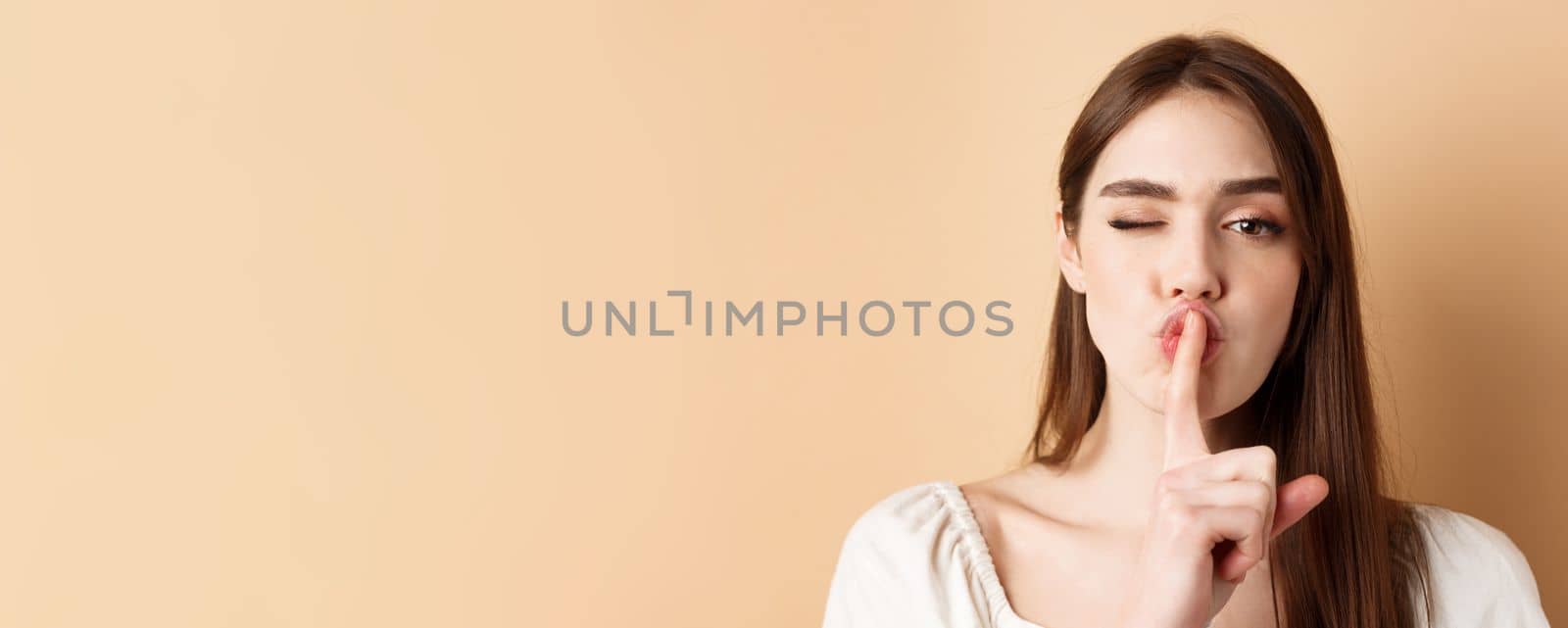 Close up of attractive woman shushing at camera, hiding secret, making hush sign and winking, show taboo sign, standing on beige background.