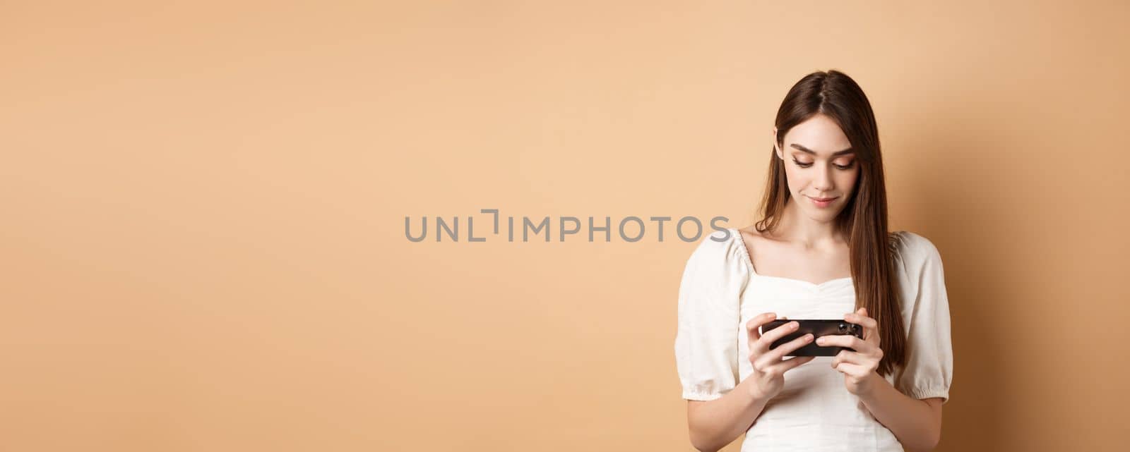 Pretty girl watching videos on smartphone, holding mobile phone horizontally and looking at screen, standing on beige background.