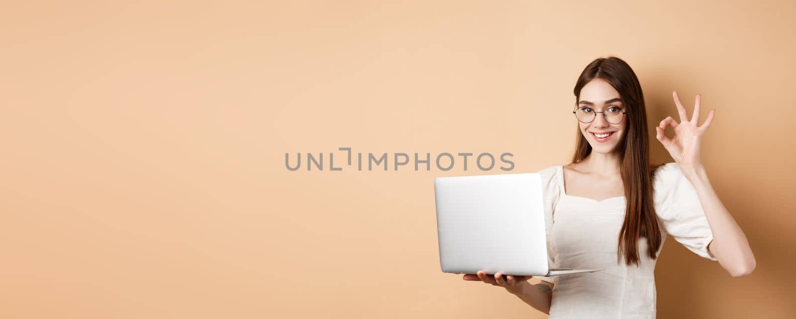 Freelance girl in glasses showing okay sign and working on laptop, guarantee good quality, recommend something online, standing on beige background by Benzoix