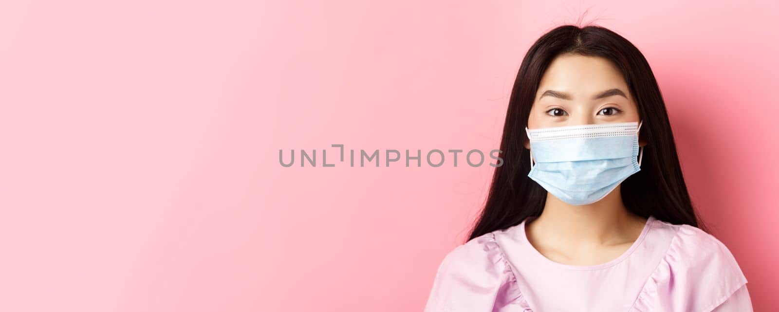 Covid-19 and healthy people concept. Close-up of asian cute girl wearing medical mask during virus outbreak, standing against pink background.