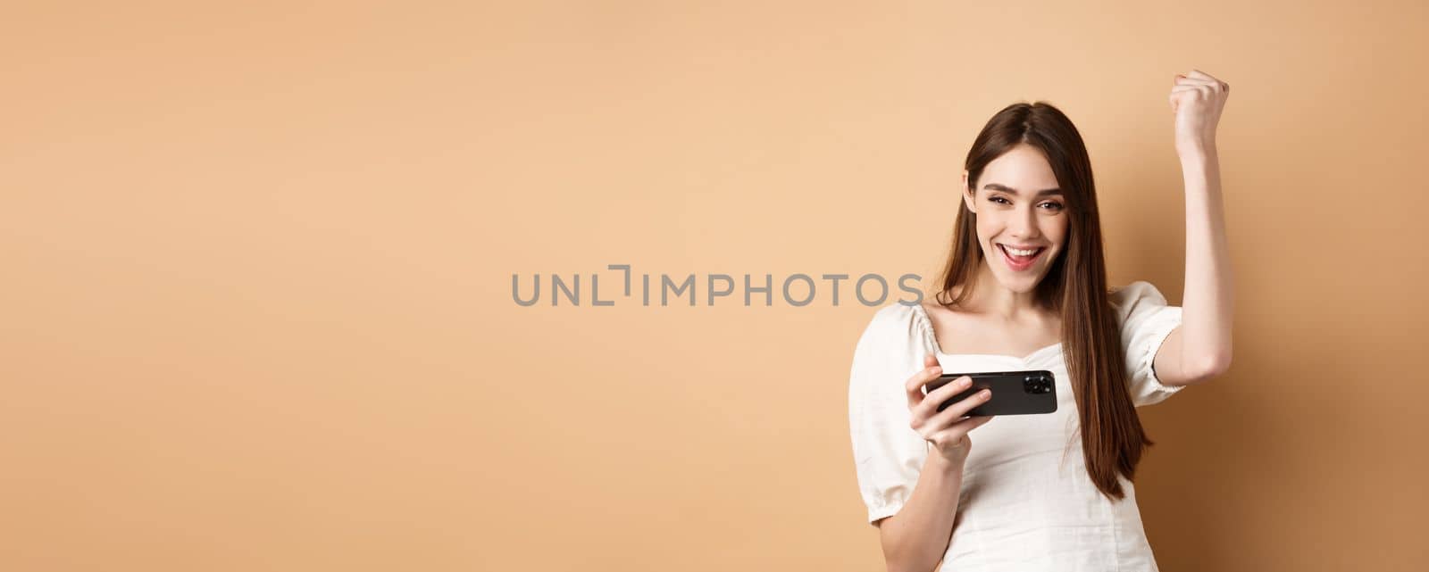Girl winning on mobile phone. Happy woman raising hand up and scream yes with joy, achieve goal in smartphone app, standing on beige background by Benzoix