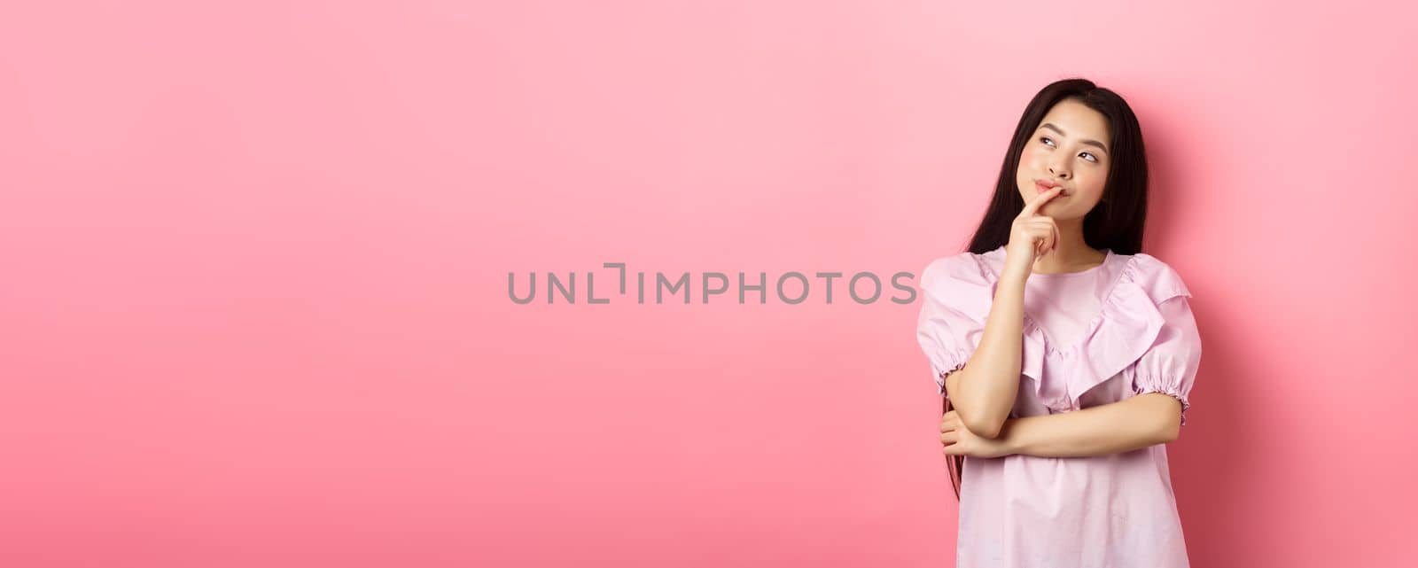 Pensive smiling asian girl making choice, looking at upper left corner logo and thinking, having an idea, standing cunning on pink background by Benzoix