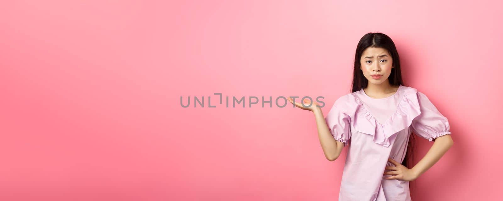 So what, who cares. Indifferent and skeptical asian girl raising arm and look unbothered at camera, dont understand whats wrong, standing against pink background.