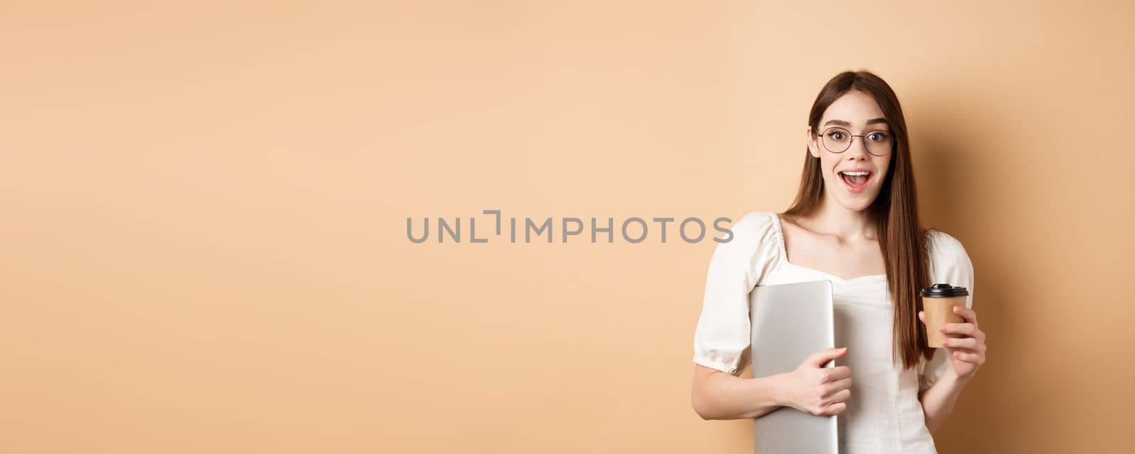 Excited woman in glasses drinking coffee from takeout cup, holding laptop and smiling happy, standing on beige background.