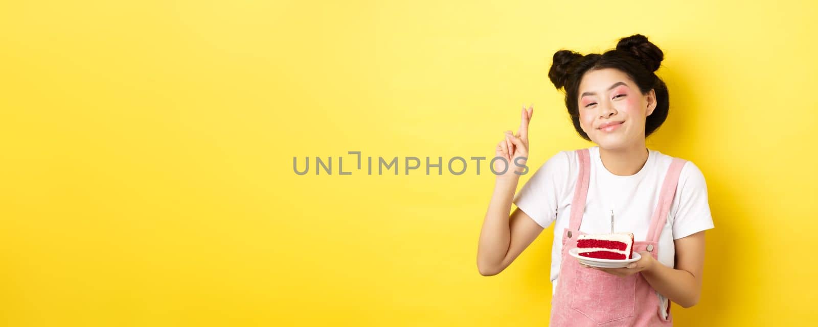 Holidays and celebration. Positive asian birthday girl cross fingers, making wish with b-day cake and lit candle, smiling happy at camera, yellow background by Benzoix