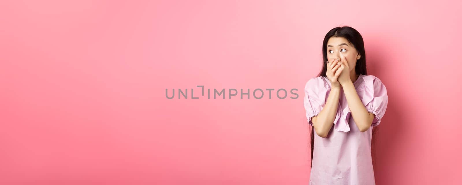 Shocked and startled asian woman looking aside at logo, covering mouth with hands speechless, standing in dress on pink background by Benzoix