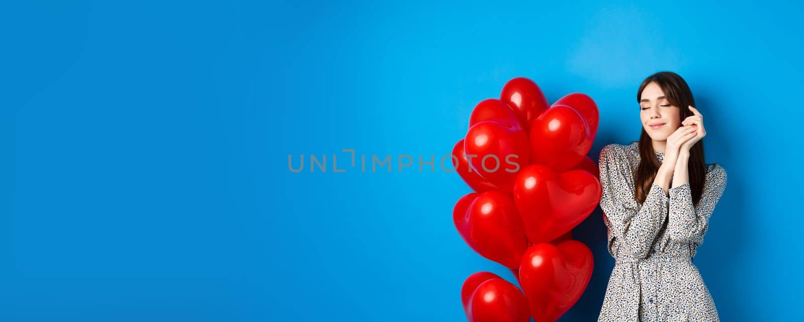 Valentines day. Dreamy romantic woman close eyes and imaging lovely date, standing near heart balloons and smiling, blue background by Benzoix