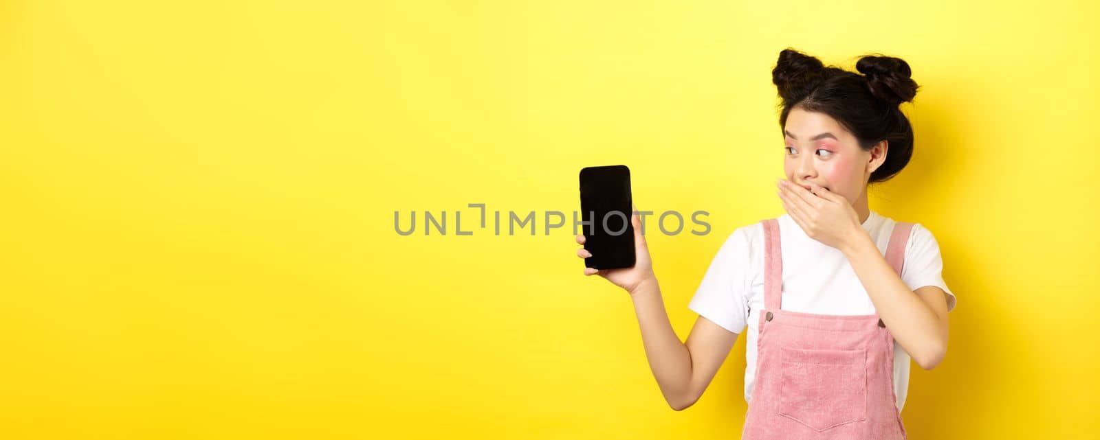 Online shopping concept. Silly japanese girl with beauty makeup, cover mouth with hand laughing and showing empty smartphone screen, show funny thing on phone, yellow background by Benzoix