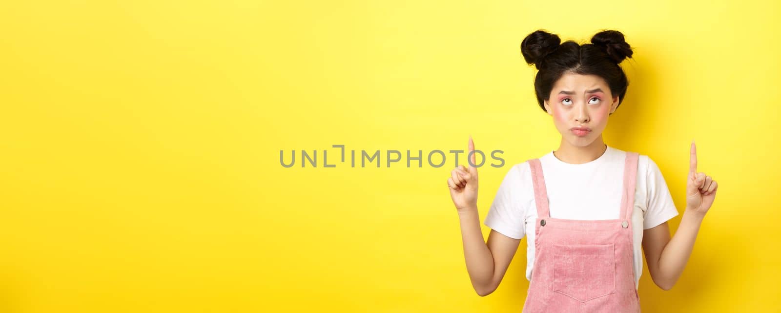 Skeptical teenage asian girl with glam pink makeup, pointing and looking up unamused, standing reluctant on yellow background by Benzoix