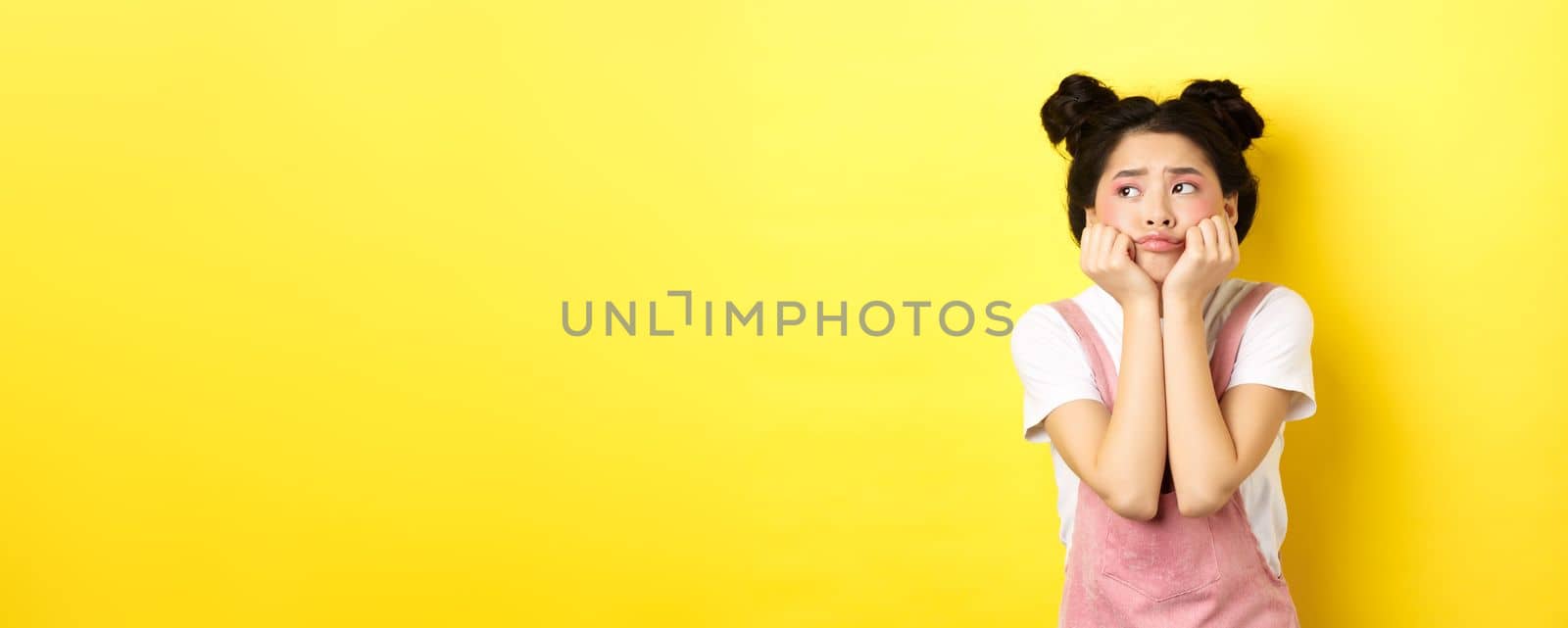 Sad moody asian girl, lean face on hands, looking left with sulking expression, standing in summer clothes on yellow background by Benzoix