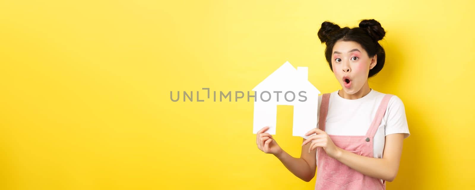 Real estate and family concept. Excited asian teen girl showing paper house cutout and gasping amazed, standing on yellow background.