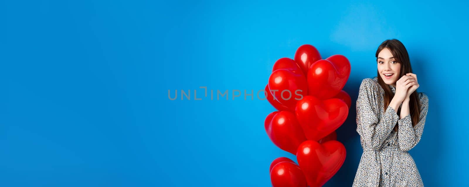 Valentines day. Lovely girlfriend ind ress smiling happy at camera, celebrating lovers holiday, standing near heart balloons, blue background by Benzoix