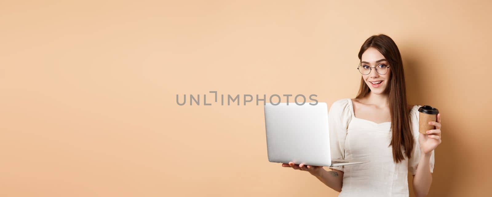 Smiling girl in glasses working on laptop and drinking coffee takeaway. Freelancer using computer at work, standing on beige background.