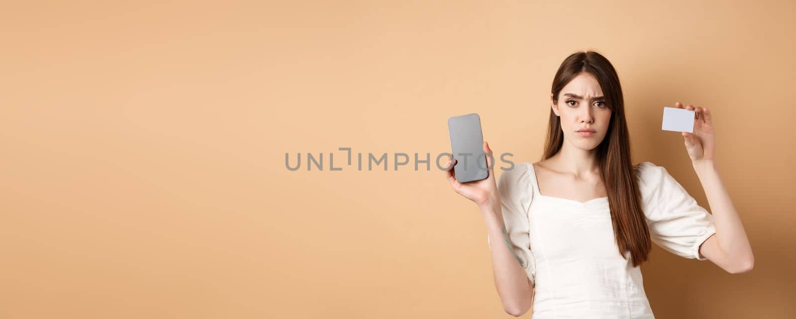 Sad and disappointed girl showing empty smartphone screen and plastic credit card, complaining and frowning, standing on beige background.