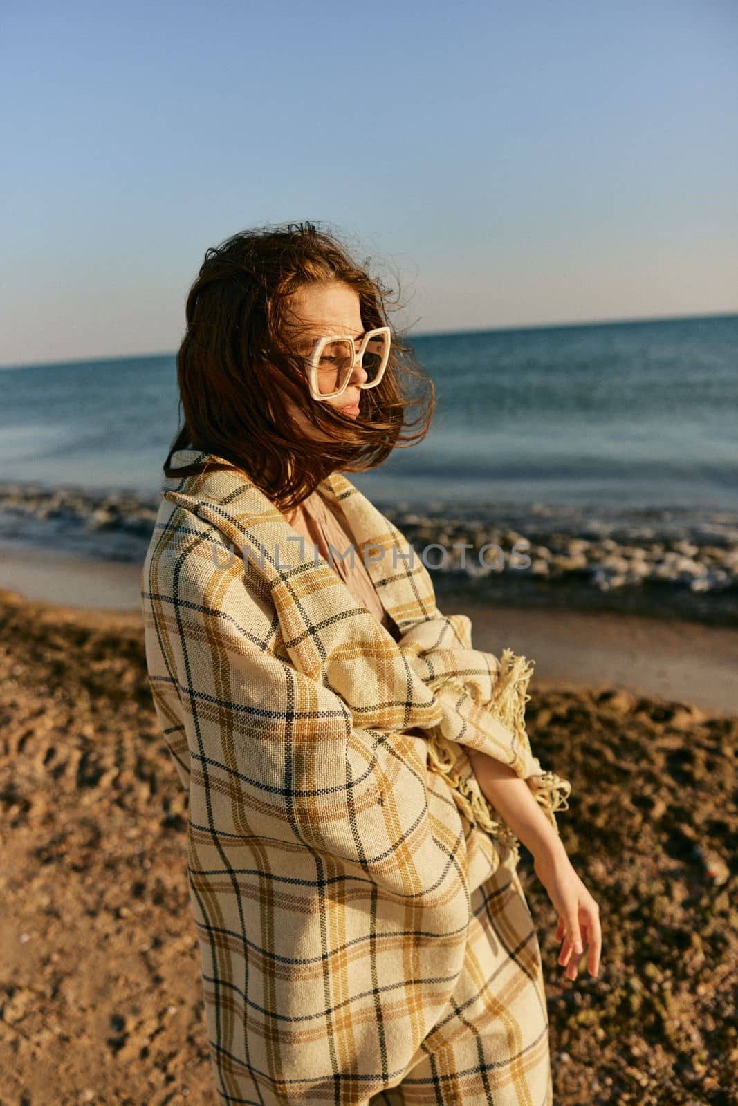 a woman in large glasses wrapped in a plaid enjoys the view of the setting sun standing on the beach by Vichizh