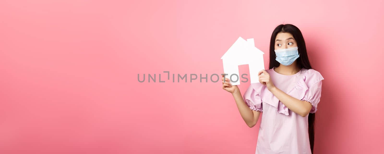 Real estate and pandemic concept. Cute asian woman in medical mask showing paper house cutout, searching for property. standing against pink background by Benzoix