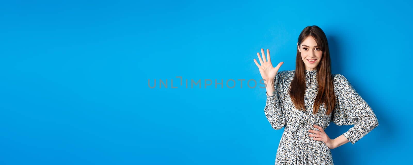 Young beautiful girl in dress with natural long hair, showing number five with fingers and smiling, standing against blue background.