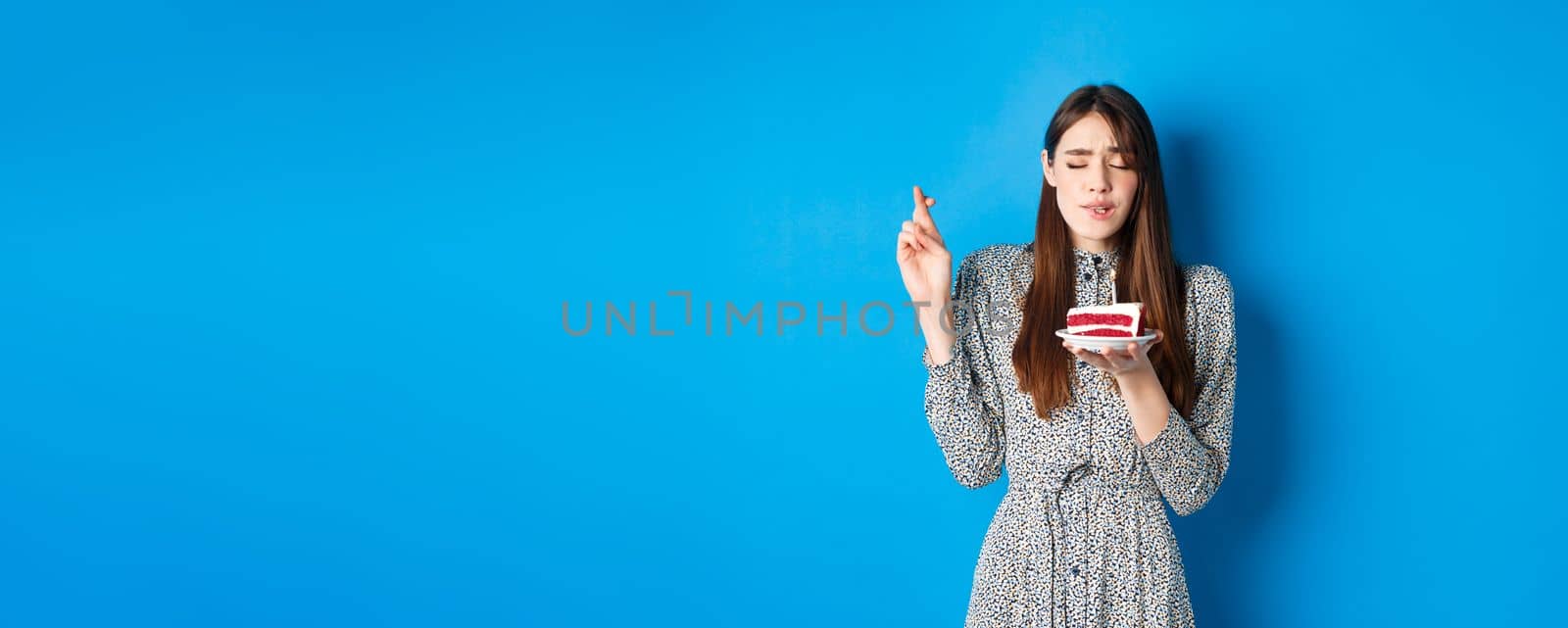 Hopeful birthday girl making wish on cake, cross fingers for good luck, standing in trendy dress against blue background.