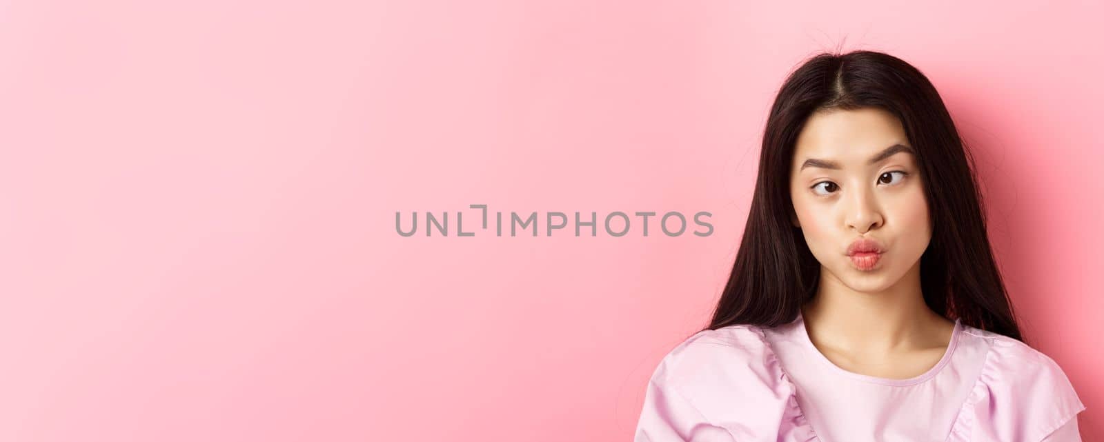 Close-up portrait of funny asian woman squinting eyes and making silly faces, standing against pink background.