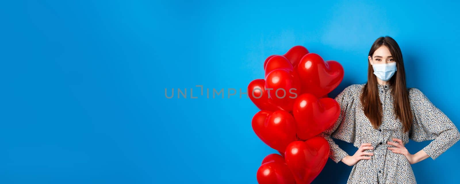Pandemic and Valentines day. Cheerful smiling girl in medical mask, standing near romantic heart balloons and looking at camera, wearing dress, blue background by Benzoix