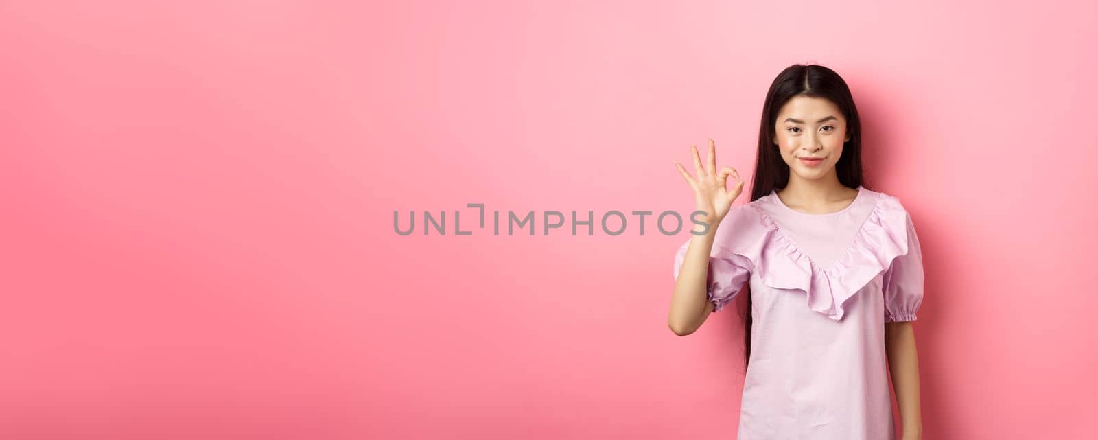 Smiling asian woman in dress showing okay sign, praise and compliment good thing, approve excellent choice, standing on pink background.