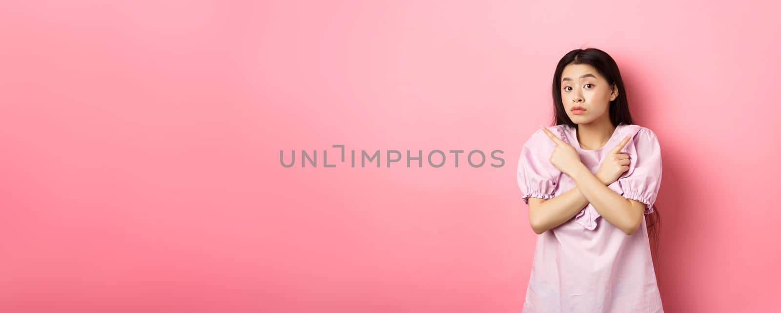 Clueless cute asian girl pointing sideways and look confused, cant make decision, standing hesitant against pink background by Benzoix